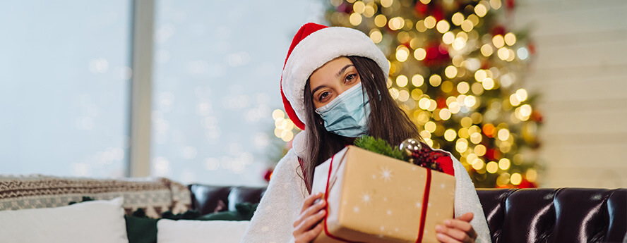 Young Women Holding Present And Wearing Medical Mask To Cover Her Face Against Coronavirus Pandemic With Christmas Lights And Tree In The Background 